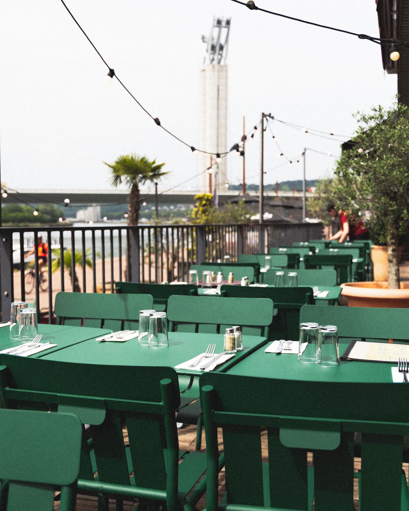 Les tables vertes dressées sur la terrasse de Toto's, guirlandes et palmier, avec la Seine et le pont Flaubert en arrière-plan